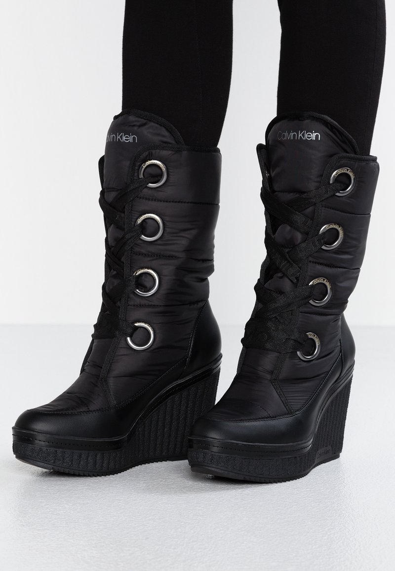 Botines negros de cuña con exterior brillante de nailon, con cordones delanteros, ojales metálicos y suela de cuña texturizada. Logo de la marca visible.