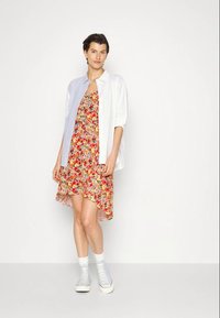Vestido floral em vermelho, amarelo e azul com uma bainha assimétrica, combinado com uma camisa branca de botões e sapatilhas brancas.