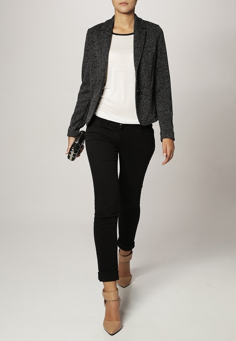 Blazer noir sur un haut blanc, associé à un jean skinny noir et des chaussures à talons hauts couleur nude, avec un tissu texturé et une coupe ajustée.