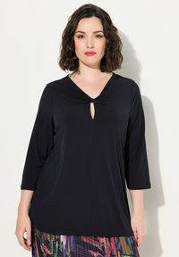 Mustatunika, jossa on V-mallinen kaula-aukko ja solmukuvio keskellä. Kolmannes hihoista ja rento istuvuus, valmistettu sileästä kankaasta.