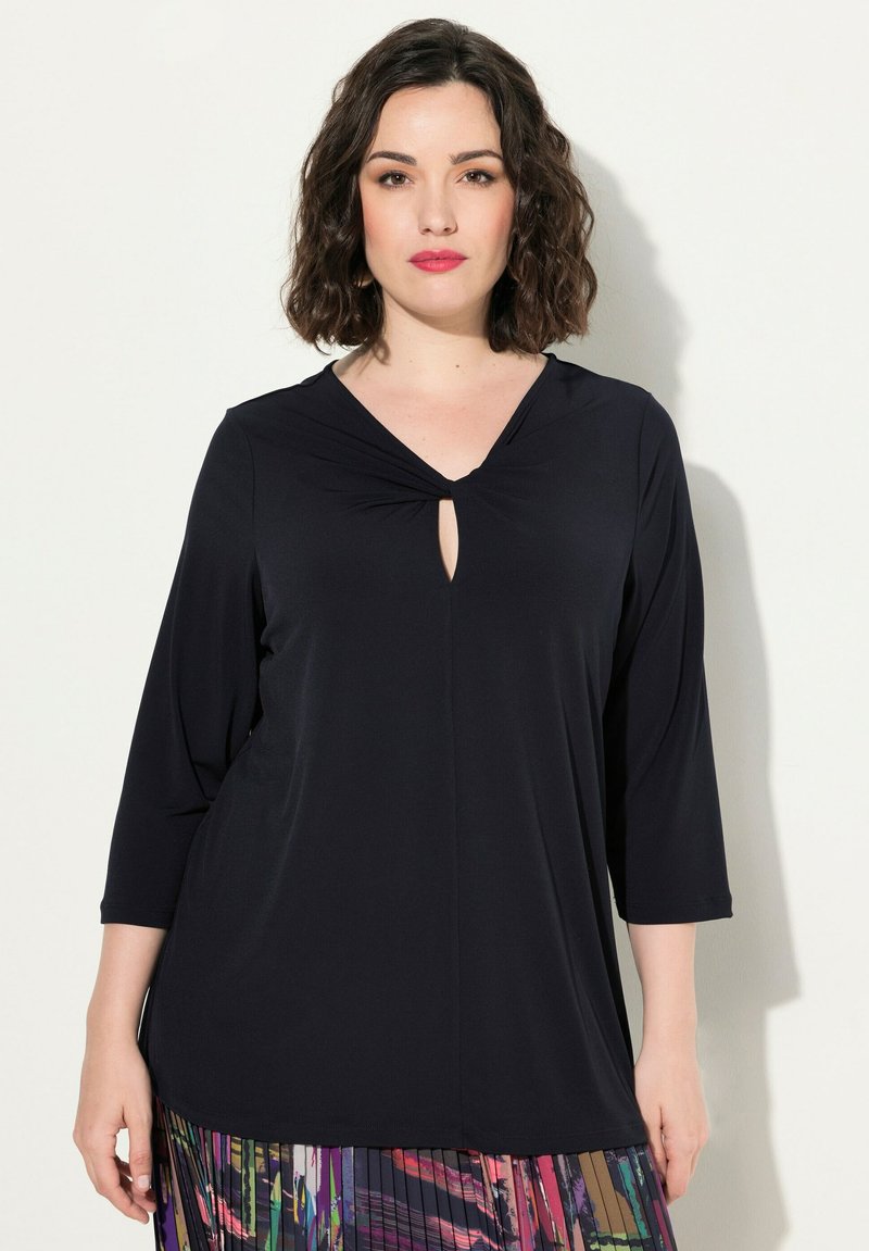 Mustatunika, jossa on V-mallinen kaula-aukko ja solmukuvio keskellä. Kolmannes hihoista ja rento istuvuus, valmistettu sileästä kankaasta.