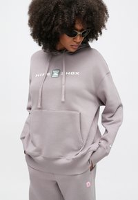 Απαλό μωβ φούτερ με κουκούλα, μπροστινή τσέπη, κορδόνια και λευκό λογότυπο "NIKE SHOX". Λείο ύφασμα, άνετη εφαρμογή, συνδυασμένο με παντελόνι στο ίδιο σχέδιο.