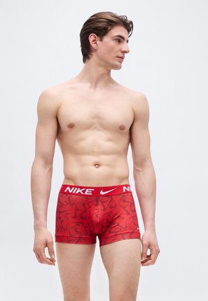 Muž bez košele s krátkymi hnedými vlasmi, oblečený v červených boxerkách Nike s čiernymi vzormi Swoosh, stojaci pred jednoduchým svetlým pozadím.