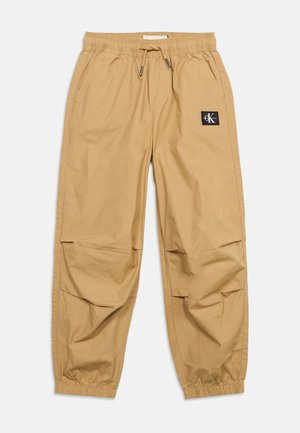 Pantalon de jogging à taille élastique couleur beige avec cordon de serrage, poches latérales, poignets froncés et petite étiquette noire avec logo sur la cuisse droite.