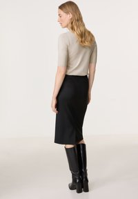 Beige kurzärmliges Oberteil, schwarze knielange Rock, schwarze kniehohe Stiefel mit Absatz. Glatte Texturen, figurschmeichelnde Silhouette, Seitenschlitz im Rock.