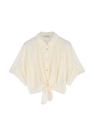 Jeune femme en pantalon rayé et chemise blanche nouée se tient dans le jardin, tenant un tuyau jaune, se protégeant les yeux de la lumière du soleil.