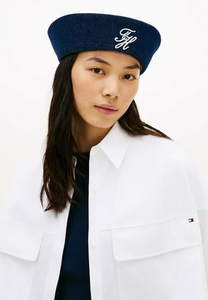 Jonge vrouw met lang donker haar draagt een marineblauwe baret met witte initialen en een witte jas over een marineblauwe top tegen een witte achtergrond.