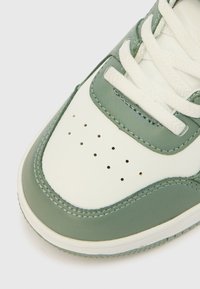 Λευκό και πράσινο αθλητικό παπούτσι με λεία δερμάτινη επιφάνεια, διακοσμημένο με διάτρητα σημεία στο μπροστινό μέρος, επίπεδα λευκά κορδόνια και ορατές ραφές.