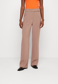 Breda byxor i ett ljust brunt tyg, med en slät front, veck och en mjuk textur. Matchat med svarta slip-on skor.