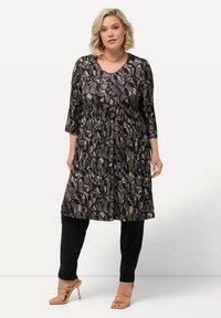 Zwarte tuniek met beige abstracte bladerenpatronen, driekwart mouwen, geplooide taille, gecombineerd met zwarte leggings en naaktkleurige sandalen met banden.