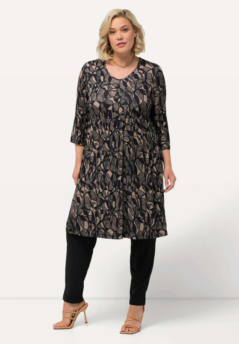 Zwarte tuniek met beige abstracte bladerenpatronen, driekwart mouwen, geplooide taille, gecombineerd met zwarte leggings en naaktkleurige sandalen met banden.