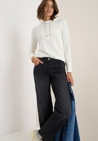 Ljusgrå prickig hoodie med dragsnören, tillsammans med svarta vida byxor. Modellen håller en blå denimjacka och har på sig ljusa sneakers.