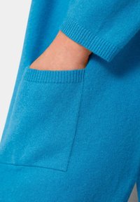 Blaues Strickkleidungsstück mit geripptem Bündchen und einer Seiten Tasche. Die Textur wirkt weich und glatt, mit einer einheitlichen Farbe und klaren Linien.