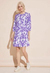 Lila-weiße gemusterte Kleid mit Dreiviertelärmeln, gestuftem Rock, rundem Ausschnitt, getragen mit weißen Turnschuhen. Glatte Stofftextur.