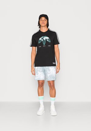 T-shirt nera con stampa grafica, dettagli bianchi e verdi, abbinata a pantaloni corti tie-dye blu chiaro, sneakers bianche e calze a righe.