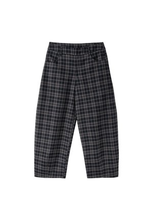 Femme aux longs cheveux portant des lunettes de soleil noires, une veste à manches courtes à carreaux, un pantalon large assorti et des chaussures blanches pointues sur fond rose.