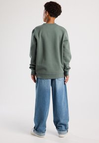 Sudadera de forro polar verde con mangas largas, puños acanalados y dobladillo. Llevada sobre unos vaqueros de denim azul claro, holgados y con los puños doblados.