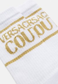 Fehér bordázott zoknik arany csillámmal írt "VERSACE COUTURE" felirattal a tetején.