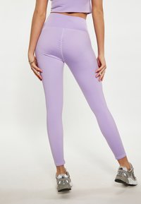 Leggings viola realizzati in un tessuto elasticizzato con una vita alta, caratterizzati da un delicato dettaglio arricciato sul retro. Abbinati a sneakers grigie e argentate.