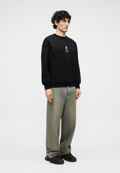 Νεαρός με σγουρά μαλλιά στέκεται φορώντας μαύρο φούτερ με μικρό λογότυπο στο κέντρο, χαλαρά γκρι τζιν και μαύρα αθλητικά παπούτσια σε απλό φόντο.