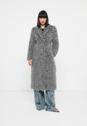 Long manteau gris en matériau texturé et duveteux, avec un col cranté et des boutons à l'avant, associé à un pantalon en denim à jambe large et des chaussures à bouts ouverts.