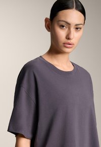 Mladá žena s tmavými vlasmi zviazanými dozadu, oblečená v voľnej tmavošedej tričku s krátkym rukávom, pozerá priamo do kamery s neutrálnym výrazom.