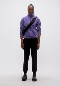 Γυναίκα που στέκεται, φορώντας μοβ φλις μπουφάν, μαύρο παντελόνι, τσάντα χιαστί και παπούτσια για πεζοπορία, μπροστά σε απλό λευκό φόντο.