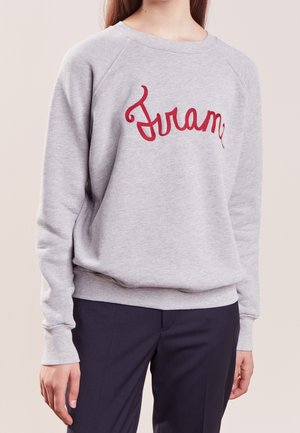 Personne portant un sweat-shirt gris clair avec un texte rouge en écriture cursive "Frame" sur la poitrine, assorti à un pantalon foncé sur fond uni.
