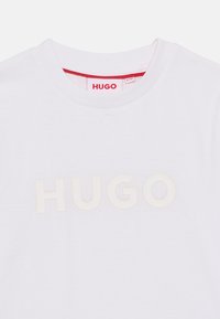 Λευκό βαμβακερό T-shirt με στρογγυλή λαιμόκοψη, με ανάγλυφο, μεγάλο κρεμ λογότυπο "HUGO" εμπρός και κόκκινη λεπτομέρεια στο εσωτερικό της λαιμόκοψης.