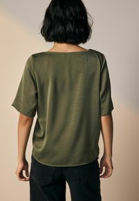 Olijfgroene blouse met korte mouwen van een gladde, licht gestructureerde stof. Heeft een brede halslijn en een afgeronde zoom, gedragen met zwarte broek.