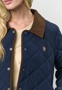 Mėlynas siūtas švarkas su rudomis diržinėmis apykaklėmis, užsegimais su snapu ir išsiuvinėtu logotipu kairėje pusėje. Matoma tekstūruota medžiaga ir raštas.