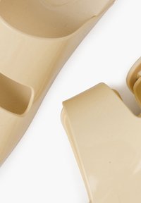 Beige plastic slip-on clogs with an open back, smooth texture, and curved shape; features a distinct, minimalist design without embellishments.