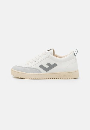 Zapatilla casual blanca de caña baja con suela beige, puntera de ante gris y logo lateral gris, con cierre de cordones y lengüeta en el talón.