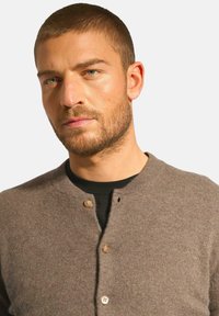 Hnědý pletený cardigan s kulatým výstřihem, který má dva viditelné knoflíky a malou dírku na přední straně. Měkká textura, volný design.