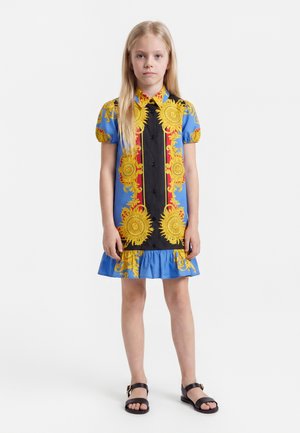 Jeune fille aux longs cheveux blonds portant une robe bleue avec un soleil jaune et des motifs baroques rouges ainsi que des sandales noires, debout sur un fond blanc.