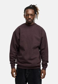 Dark purple sweatshirt with a high neckline, loose fit, and ribbed cuffs. Worn with dark jeans, featuring a smooth texture.