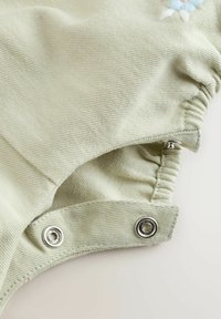 Tissu vert clair avec bords élastiques et boutons-pression argentés, montrant un gros plan détaillé de la fixation sur un matériau côtelé doux.