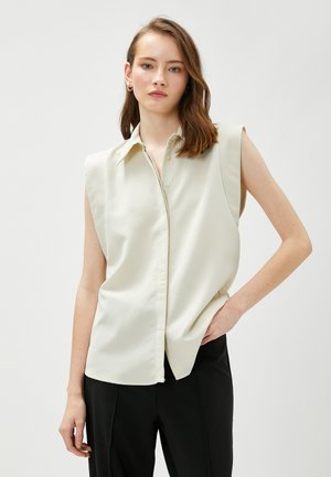 Blusa senza maniche color beige chiaro con colletto, chiusura frontale con bottoni e una trama liscia. Abbinata a pantaloni neri.