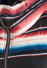 Vaatteena on pehmeä takki, jossa on vetoketju. Se esittelee moniväristä raita-kuviota punaisen, sinisen ja valkoisen sävyissä mustaa taustaa vasten. Pintamateriaali on sileä.