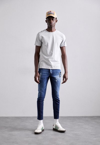 T-shirt en coton gris, jeans skinny déchirés bleus avec une poche latérale, chaussettes blanches et baskets blanches avec des rayures vertes. Casquette jaune sur la tête.