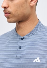 Νέος άνδρας που φοράει μπλε πουκάμισο με κουμπιά και καρό σχέδιο, με λευκό λογότυπο Adidas στο στήθος.