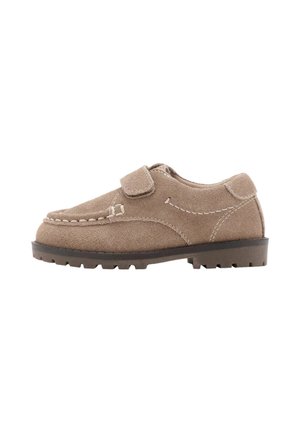 Zapato de ante beige único con tira de velcro, costuras blancas y suela gruesa y acanalada de color oscuro, orientado hacia la izquierda sobre un fondo blanco.
