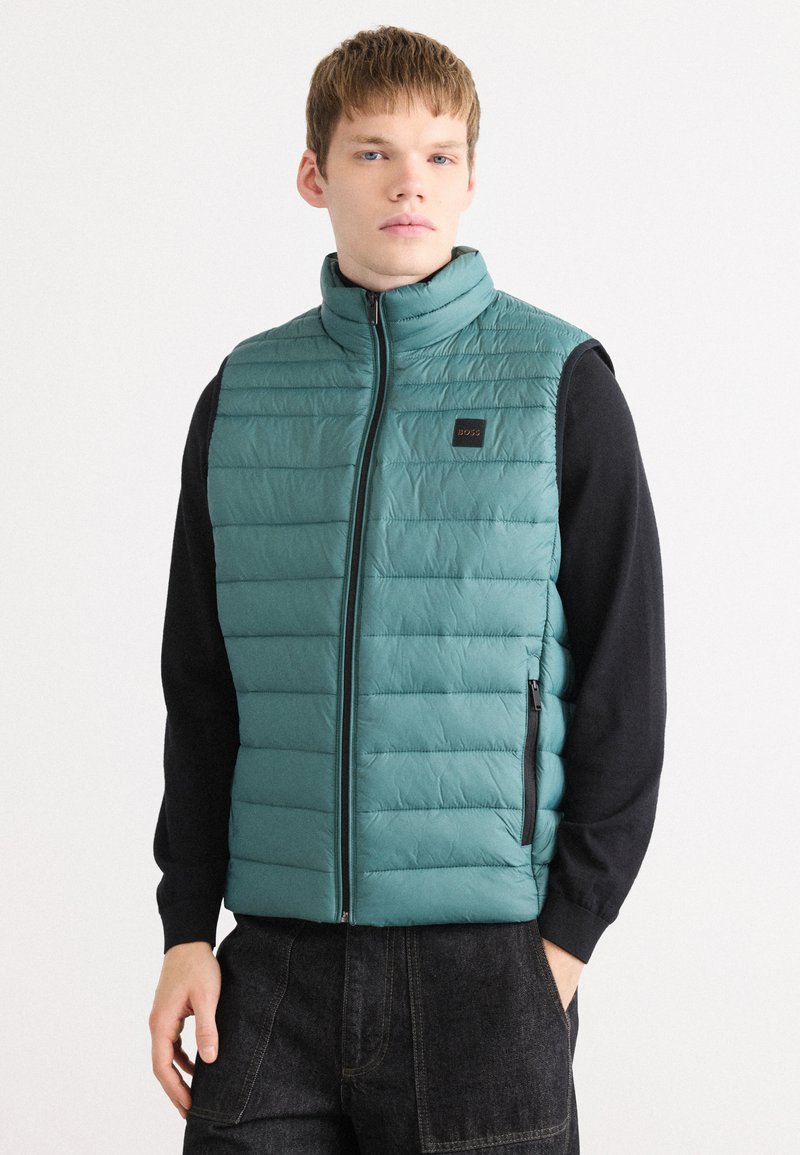 Gilet imbottito in teal, con design a cuciture orizzontali, colletto alto e tasche laterali con zip. Accenti neri alle aperture per le braccia.