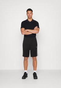 Polo noir associé à un short noir avec des accents de panneau latéral gris. Modèle portant des baskets noires et des chaussettes blanches, les bras croisés.