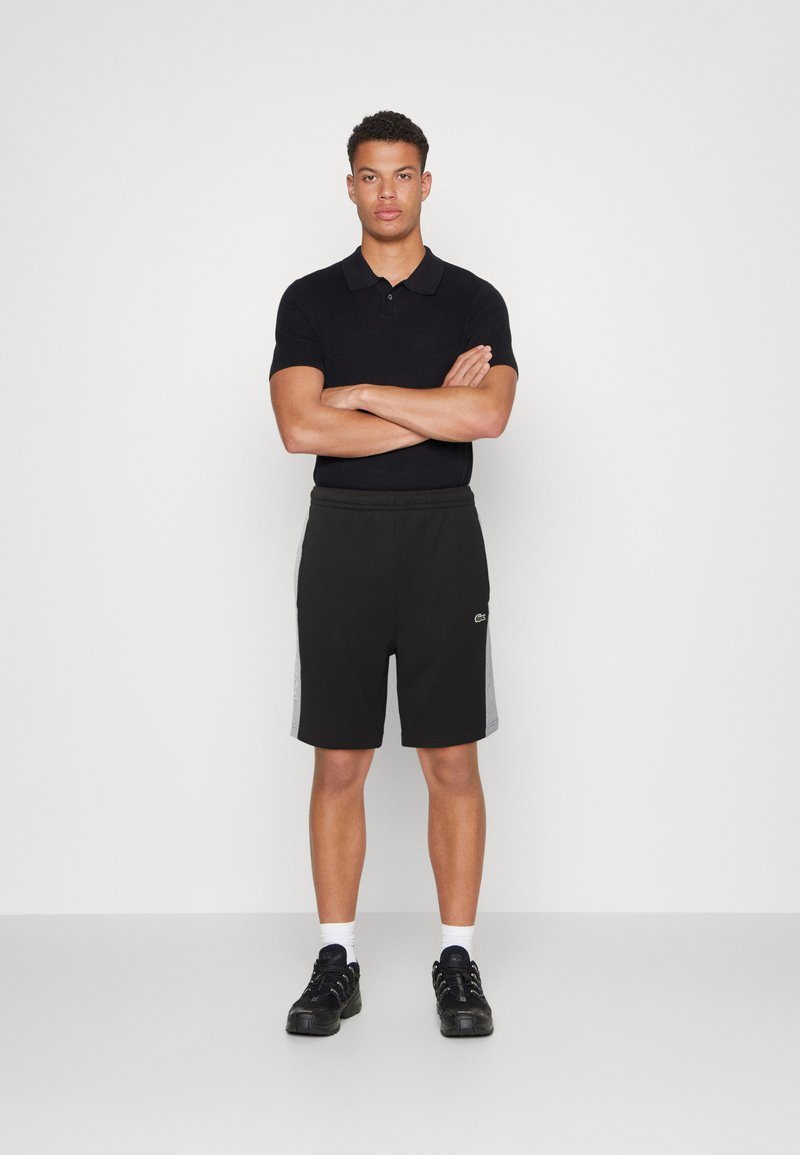 Polo noir associé à un short noir avec des accents de panneau latéral gris. Modèle portant des baskets noires et des chaussettes blanches, les bras croisés.