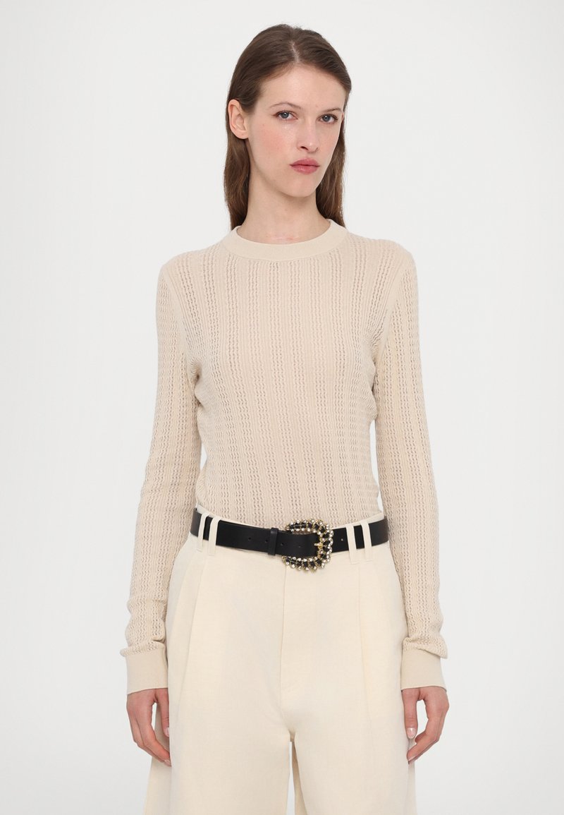 Beżowy sweter dzianinowy o prążkowanym wzorze, okrągłym dekolcie i długich rękawach, zestawiony z kremowymi spodniami z wysokim stanem i czarnym paskiem z ozdobami.