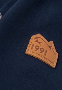 Navy fabric features a prominent brown leather tag with embossed text "1991 SWEDEMOUNT" and mountain graphics.