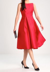 Robe A-ligne en satin rouge avec un design sans manches, jupe plissée et escarpins noirs, tenant une pochette noire.