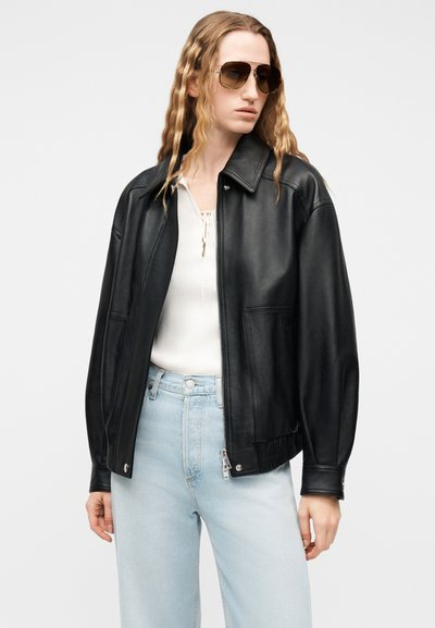 Νεαρό άτομο με μακριά κυματιστά μαλλιά που φοράει σκουρόχρωμα γυαλιά ηλίου, μαύρο δερμάτινο μπουφάν πάνω από λευκό τοπ και ανοιχτό μπλε τζιν με ψηλή μέση.