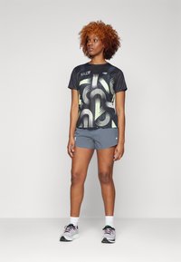 Svart trenings t-skjorte med grønt abstrakt mønster, paret med grå shorts. Modellen har på seg hvite og svarte joggesko med grønne detaljer.